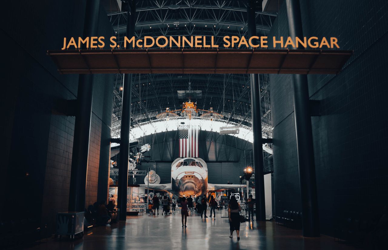 Interior view of the James S. McDonnell Space Hangar featuring a showcased spacecraft with visitors exploring.